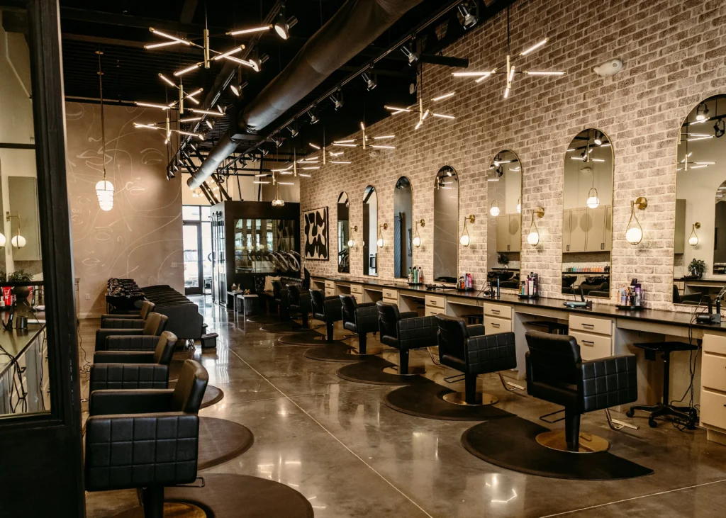 Modern luxury hair salon interior with styling stations, arched mirrors, black salon chairs, exposed brick walls, and contemporary lighting. The Beauty Vault at New Riverside Village's interior was built by Trulane General Contracting, Inc.